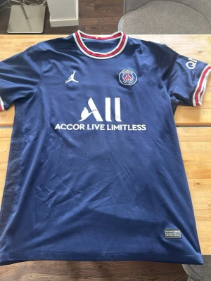 PSG matchtröja blå Jordan - Snygg blå Paris Saint-Germain matchtröja från Jordan med klubbmärke på bröstet och vit logga för Accor Live Limitless. Tröjan har rund hals med röd, vit och blå kant samt matchande detaljer på ärmarna. Perfekt för fotboll och sport.