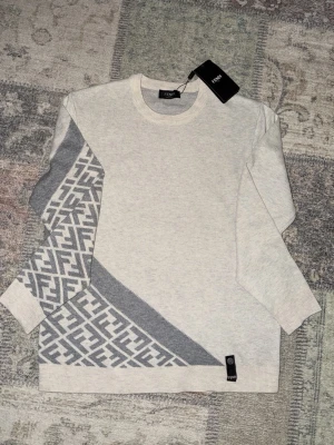 Beige tröja med grått mönster från Fendi - Snygg beige tröja från Fendi med grått grafiskt mönster på ena ärmen och nedre delen av tröjan. Rund halsringning och långa ärmar. Perfekt för dig som gillar exklusiva detaljer och stilren design. Materialet känns mjukt och bekvämt.