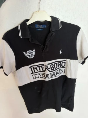 Ralph Lauren Piké / Tshirt S - Säljer en svartvit t-shirt från Polo Ralph Lauren med klassisk krage och knappar. T-shirten har tryck med texten 'INTER-BORO CYCLE SERIES' och broderad logga på bröstet. Tillverkad i bomull och har korta ärmar., Storlek S