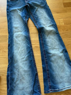 Blå bootcut jeans från Gina Tricot - Säljer ett par blå bootcut jeans från Gina Tricot med snygga slitningar och kontrastsömmar i rött. Jeansen har klassisk femficksmodell och är tillverkade i denim med skön stretch. Perfekta för dig som gillar en avslappnad men trendig look. Lowwaist