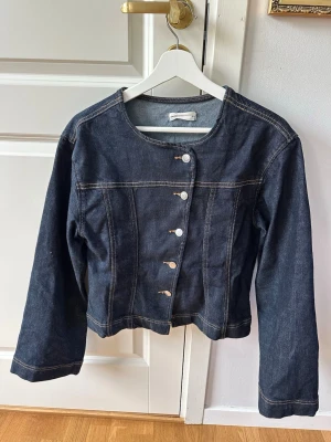 Mörkblå jeansjacka med rund hals - Snygg mörkblå jeansjacka med rund halsringning och asymmetrisk knäppning framtill. Jackan har långa ärmar och kontrastsömmar som ger en cool look. Passar perfekt till ett par jeans eller kjol för en avslappnad stil.