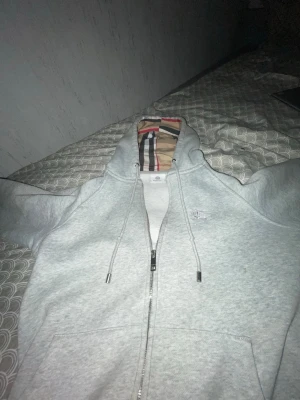 Ljusgrå zip-hoodie från Burberry - Snygg ljusgrå hoodie med dragkedja från Burberry. Klassisk huva med Burberrys rutiga mönster på insidan och broderad logga på bröstet. Mjukt material och dragsko med metalländar där det står Burberry. Perfekt för en stilren och avslappnad look.