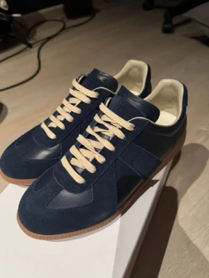 Blå Maison Margiela Replica sneakers - Snygga blå Maison Margiela Replica sneakers i mocka och läder med beige snören och gummisula. Klassisk låg modell med diskret logga på plösen och vit insida. Perfekta för dig som gillar stilrena och tidlösa sneakers.
