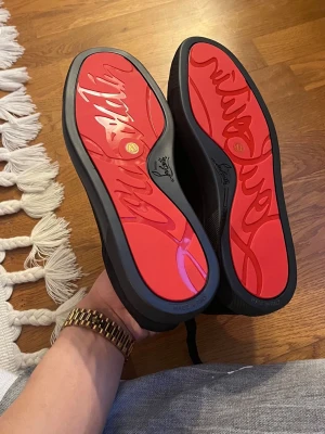 Svarta Louboutin sneakers - Säljer ett par svarta sneakers från Christian Louboutin med ikonisk röd sula och diskret logga på sidan. Skorna är låga och har ovandel i mocka och läder, samt svarta skosnören. Perfekta för dig som vill sticka ut med stilrena detaljer.
