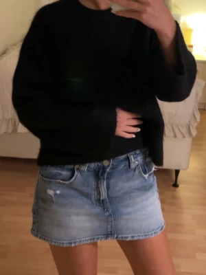 Blå jeanskjol med slitningar - Säljer en klassisk blå jeanskjol i kort modell med slitna detaljer och rå kant. Kjolen har fem fickor, bälteshällor och stängs med knapp och dragkedja. Perfekt för en avslappnad och trendig look.