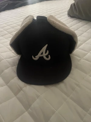 Svart New Era keps med öronlappar - Säljer en svart keps från New Era med Atlanta Braves-logga framtill och vita öronlappar i mjukt teddy-material. Kepsen har böjd skärm och klassisk passform, perfekt för kyligare dagar. Snygg detalj med broderad New Era-logga på sidan.