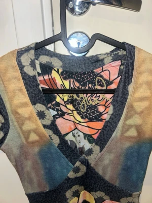 Färgglad klänning från Desigual - Unik kortklänning från Desigual med färgstarkt blommönster i orange, blått, beige och svart. Klänningen har v-ringning, korta ärmar och mjukt material som känns skönt mot huden. Perfekt för dig som gillar statement-plagg och kreativa prints.