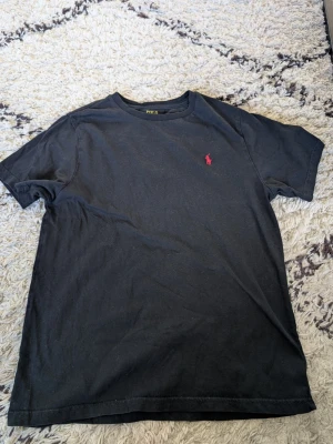 Svart t-shirt från Polo Ralph Lauren stl 14-16 - Klassisk svart t-shirt från Polo Ralph Lauren med den ikoniska röda logon broderad på bröstet. T-shirten har rund halsringning och korta ärmar. Perfekt basplagg som funkar till allt och är tillverkad i mjuk bomull.