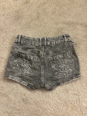 Grå denimshorts med broderade fickor - Säljer ett par grå denimshorts med dubbla knappar framtill. Shortsen har två bakfickor med vita broderade hjärtan och snygga slitningar för en cool look. Perfekta för sommaren och har bälteshällor för extra stylingmöjligheter.