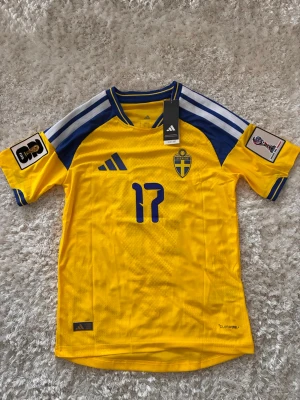 Svensk landslagströja Adidas gul - Gul svensk landslagströja från Adidas med blå detaljer, nummer 17 på bröstet och blått landslagsemblem. Tröjan har vita och blå ränder på axlarna, FIFA World Cup-märke på ärmen och är tillverkad i lätt funktionsmaterial.
