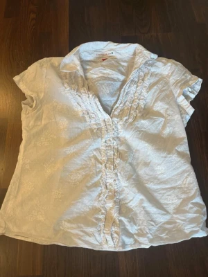 Vit blus med volangdetaljer - Snygg vit blus med korta ärmar och volangdetaljer längs knäppningen framtill. Blusen har en klassisk krage och diskret blommönster i tyget. Perfekt för en stilren och fräsch look.