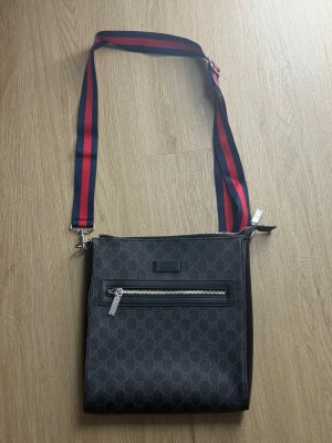 Guccci axelväska med monogram och röd-blå rem - Snygg axelväska från Guccci i svart med klassiskt GG-monogram. Väskan har en justerbar axelrem i rött och blått, silverfärgade dragkedjor och en praktisk ytterficka. Materialet är canvas med läderdetaljer. Perfekt för dig som gillar stilrena accessoarer.