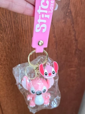 Rosa Stitch nyckelring med figur - Säljer en söt rosa Stitch-nyckelring med en mjuk silikonrem där det står 'Stitch Love'. Nyckelringen har en 3D-figur av Stitch i rosa och en liten matchande charm. Perfekt att hänga på väskan eller nycklarna för en gullig touch.