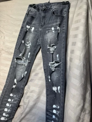 Grå slitna skinny jeans med hål - Säljer ett par grå skinny jeans med rejält slitna detaljer och stora hål på framsidan. Jeansen har en tvättad look, fem fickor och vita färgstänk för extra edge. Perfekta för dig som gillar streetstyle och vill sticka ut. (Pris kan diskuteras)