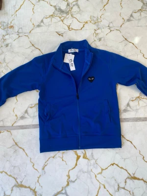Blå zip-hoodie från Comme des Garçons - Snygg blå zip-hoodie från Comme des Garçons PLAY med svart hjärta-logga på bröstet. Tröjan har dragkedja framtill, två fickor och ribbade muddar. Perfekt för en chill och stilren look.