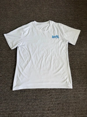 Vit t-shirt  - Vit t-shirt med blått 'AMOR'-tryck på bröstet. Klassisk rund halsringning och korta ärmar. Tillverkad i mjuk bomull för en skön känsla. Enkel och stilren design som passar till det mesta.
