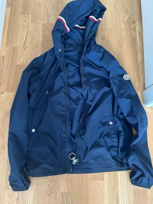 Marinblå vindjacka från Moncler - Snygg marinblå vindjacka från Moncler med huva och dragkedja.  den är storlek. s och m det är inga problem med den dragkedjan funkar som vanligt och jag har byten och oris kan alltid diskuteras den är mörkblå 