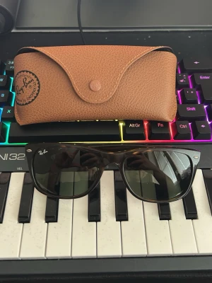 Ray-Ban Wayfarer solglasögon brun - Klassiska Ray-Ban Wayfarer solglasögon med mörkbruna bågar och svarta glas. Snygg retroform som passar till de flesta stilar. Kommer med original Ray-Ban fodral i brunt skinn. Perfekta för soliga dagar!
