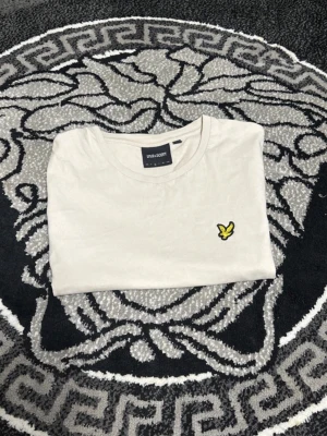 Vit t-shirt från Lyle & Scott - Klassisk vit t-shirt från Lyle & Scott med rund hals och korta ärmar. På bröstet sitter den ikoniska gula fågel-loggan broderad. Tillverkad i mjuk bomull för en skön och avslappnad stil.