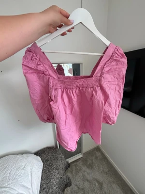 Rosa puffärm topp med smock - Söt rosa topp med puffärmar och smockad kant vid halsen. Toppen har en fyrkantig ringning och är gjord i ett lätt bomullstyg. Perfekt för dig som gillar en feminin och trendig stil.