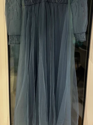 Ljusblå långklänning med paljetter - Magisk ljusblå långklänning från Ever Pretty med transparenta, långärmade meshärmar och v-ringning. Överdelen är dekorerad med glittriga paljetter och kjolen är i flera lager tyll för en drömmig look. Perfekt för dig som vill sticka ut.