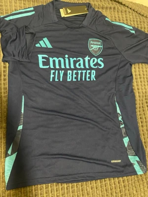 Arsenal träningströja Adidas blå - Mörkblå Arsenal träningströja från Adidas med ljusblå detaljer och logga. Tröjan har trycket 'Emirates Fly Better' på bröstet och är tillverkad i lätt och ventilerande Aeroready-material. Perfekt för fotbollsträning eller sport.