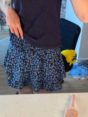 Blommig kortkjol i blått och svart - Söt kortkjol med blått och svart blommönster. Kjolen har en volangig nederdel och resår i midjan för skön passform. Perfekt för dig som gillar en lekfull och trendig stil.