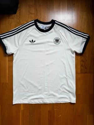 Tyskland adidas tshirt - Säljer en klassisk vit Adidas T-shirt med svarta ränder på axlarna, svart krage och svarta muddar. T-shirten har Adidas-logga och ett tyskt landslagsemblem på bröstet. Perfekt för dig som gillar retrostil och sportig look.