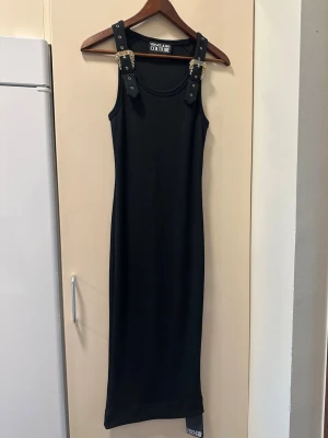 Svart långklänning från Versace Jeans Couture - Versace Jeans Couture klänning – Ny med tagg Elegant svart klänning från Versace Jeans Couture i ribbad stretchkvalitet med exklusiva guldfärgade detaljer och ikoniska spännen vid axlarna. En tidlös modell som passar lika bra till fest som till en mer uppklädd vardagslook. ✔ Ny med originaltagg kvar ✔ Exklusiva Versace-detaljer ✔ Figurnära och smickrande passform ✔ Stretchigt och bekvämt material ✔ Klassisk svart färg som aldrig går ur stil Klänningen är oanvänd och i nyskick. FRAKTAR INOM 24H🩷