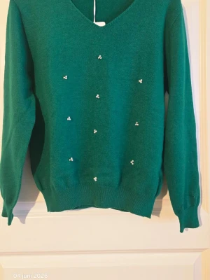 Grön stickad tröja med pärldetaljer - Snygg grön stickad tröja med v-ringning och små vita pärldetaljer framtill. Tröjan har långa ärmar och ribbade muddar vid ärmslut och nederkant. Perfekt för dig som vill ha något både stilrent och unikt.