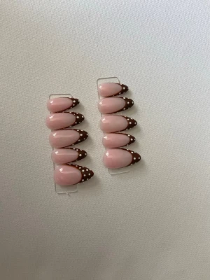 Press-on naglar rosa med bruna tippar - Säljer ett set press-on naglar i ljusrosa med bruna tippar dekorerade med vita prickar. Naglarna har en mandelformad design och ger en söt och trendig look. Perfekt för dig som vill ha snygga naglar snabbt och enkelt.