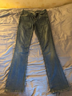 Blå bootcut jeans med låg midja - Säljer ett par klassiska blå bootcut jeans med låg midja och från Zara. Jeansen har en ljus tvätt och subtila slitningar för en avslappnad look. Tillverkade i denim med dragkedja och knapp framtill.