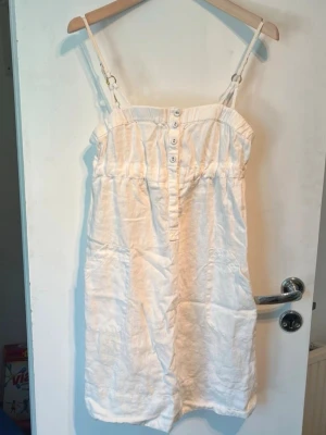 Vit Guess Jeans linneklänning med knappar  - Söt vit kort klänning med smala axelband och dekorativa guldringar. Klänningen har knappar framtill och är gjord i ett lätt material, perfekt för varma dagar. Enkel och stilren design som passar till många tillfällen.