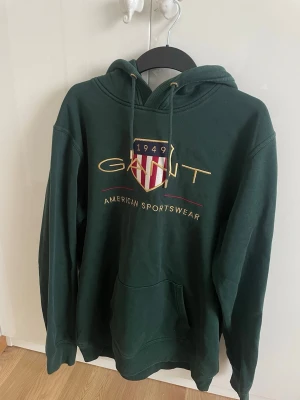 Mörkgrön hoodie från GANT 2XL - Snygg mörkgrön hoodie från GANT med broderad logga och texten 'American Sportswear' på bröstet. Klassisk känguruficka framtill, justerbar huva med snören och mjukt material. Perfekt för en avslappnad och sportig stil.