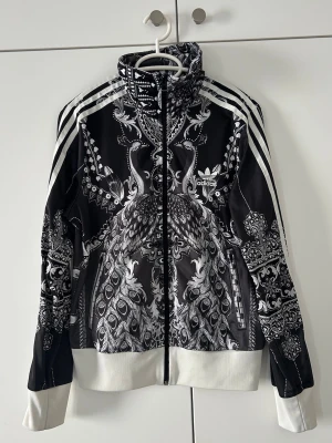 Svartvit adidas vindjacka med mönster - Unik svartvit vindjacka från adidas med detaljerat mönster av påfåglar och paisley. Jackan har dragkedja framtill, höga muddar i vitt och klassiska adidas-ränder längs ärmarna. Perfekt för dig som vill sticka ut med en sportig och cool look.
