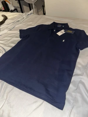 Marinblå pikétröja från Polo Ralph Lauren - Klassisk marinblå pikétröja från Polo Ralph Lauren med korta ärmar, krage och knappar framtill. Tröjan har den ikoniska broderade loggan på bröstet och är tillverkad i bomull för en bekväm känsla.