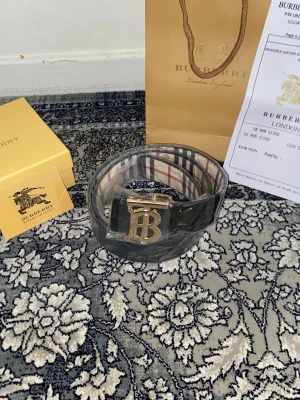 Burberry svart bälte med guldspänne - Snyggt svart bälte från Burberry med klassiskt randigt mönster på insidan och ett stort guldfärgat BB-spänne framtill. Perfekt accessoar för att lyfta din outfit med en lyxig touch. Kommer med originalförpackning och påse.
