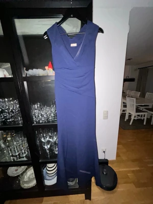 Marinblå Klänning - Elegant marinblå långklänning med v-ringad omlott framtill och ärmlös design. Klänningen har en smickrande passform och lätt drapering i midjan. Perfekt för speciella tillfällen där du vill känna dig extra fin. Köpt på bubbleroom. Säljer då den inte passade min flickvän. Skicket är otroligt bra! Pris kan diskuteras