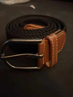 Svart flätat bälte med bruna detaljer - Stilrent svart flätat bälte med bruna skinn-detaljer och silverspänne. Perfekt accessoar för att lyfta din outfit med en klassisk touch. Bältet har en snygg kontrast mellan det svarta bandet och de bruna skinnpartierna vid spännet.