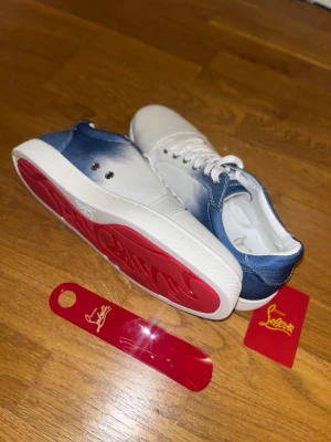 Christian Louboutin blåvita jeans modellen - Snygga sneakers från Christian Louboutin i vitt och blått tyg med klassisk röd sula. Skorna har lågt skaft, vita skosnören och metallfärgade öljetter. Perfekt för dig som vill sticka ut med exklusiva detaljer och ikonisk design.