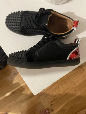 Svarta sneakers med nitar och röd detalj - Säljer ett par svarta sneakers från Christian Louboutin med coola svarta nitar på tån och röd metallic detalj på hälen. Skorna har snörning, svart sula och ovandel i läder och textil. Perfekta för dig som vill sticka ut med din stil.