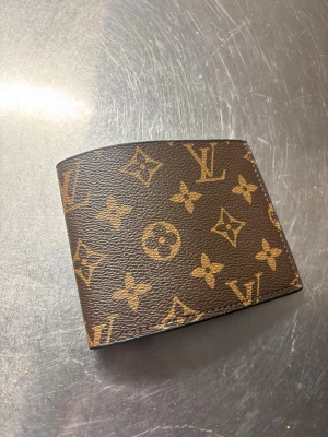 Louis Vuitton brun monogram plånbok - Snygg brun plånbok från Louis Vuitton med klassiskt monogrammönster i beige. Tillverkad i tåligt läder med flera kortfack och plats för sedlar. Perfekt accessoar för dig som gillar lyxiga detaljer och tidlös design.