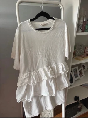 Vit kort klänning med volang - Supersöt vit kort klänning med rund halsringning och korta ärmar. Klänningen har dubbla volangdetaljer framtill som ger en lekfull och trendig look. Perfekt för dig som gillar en avslappnad men ändå stylish stil.