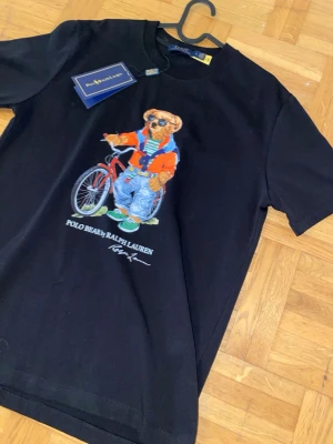 Svart Polo Bear t-shirt från Ralph Lauren - Svart t-shirt från Ralph Lauren med ikoniskt Polo Bear-tryck framtill där björnen har solglasögon och cykel. Klassisk passform och rund hals. Tillverkad i mjuk bomull som känns skön mot huden. Perfekt för dig som vill ha en statement-tee med cool vibe.