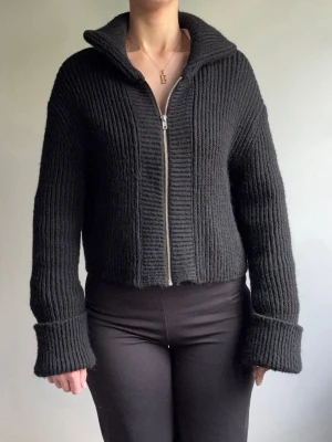Svart ribbstickad ziptröja - Säljer en svart ribbstickad tröja med dragkedja framtill och hög krage. Tröjan har långa ärmar med breda muddar och är något croppad i modellen. Perfekt för lager på lager och enkel att matcha med olika outfits.