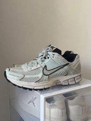 Nike vita sneakers - Nike sneakers i vitt och ljusgrått med svart swoosh. Ovandel i mesh och syntetiska paneler för extra ventilation och support. Tjock, dämpad sula med detaljer i svart och grått. Klassisk snörning och bekväm passform. Perfekta för dig som gillar sportig och trendig stil.