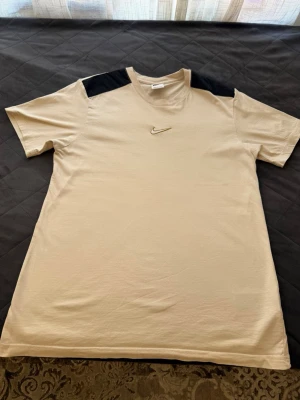 Beige Nike t-shirt med svarta detaljer - Snygg beige t-shirt från Nike med svarta partier på axlarna och klassisk Nike-logga broderad på bröstet. Tillverkad i mjuk bomull och har en normal passform med korta ärmar. Perfekt för en avslappnad och sportig stil.                           Pris går att diskutera.