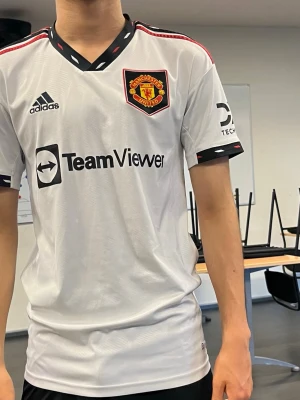 Manchester United bortatröja Adidas - Snygg vit Manchester United bortatröja från Adidas med svarta och röda detaljer. Tröjan har klubbmärke på bröstet, Adidas-logga och TeamViewer sponsortryck. Tillverkad i lätt och ventilerande material, perfekt för fotboll eller träning.