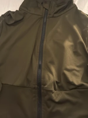 Olivgrön träningströja med dragkedja - Snygg olivgrön träningströja med hög krage och hel dragkedja framtill. Tillverkad i ett slätt, lätt material som ger en sportig look. Perfekt för dig som gillar stilrena och funktionella plagg. Klipp vid händerna för att få plats me tumme