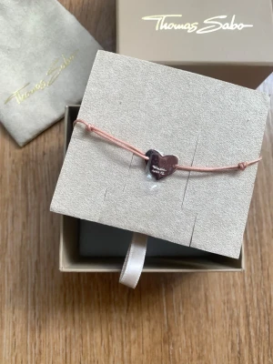Hjärtarmband från Thomas Sabo - Sött armband från Thomas Sabo med ett silverfärgat hjärta i metall på en tunn, ljusrosa textiltråd. Hjärtat har en blank finish och är graverat med märkesnamnet. Perfekt accessoar för en personlig touch.